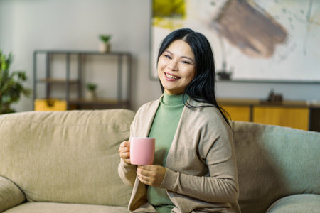 mature Asian woman enjoys a hot cup of tea while relaxing on her cozy living room sofa. High quality photoの写真素材