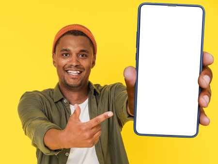 Smiling African student poses with large mobile phone featuring white blank screen, on yellow background with copy space. This image is perfect for product placement or advertising concepts. High quality photoの写真素材