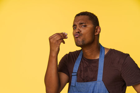 Stylish African American cook wearing blue apron sends kiss with his fingers in Italian manner, against bright yellow background with copy space for text or product placement. High quality photoの写真素材