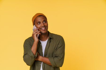 Smiling African man wearing orange cap is having phone call, with copy space on yellow background. Perfect for product placement and advertising concepts. High quality photoの写真素材