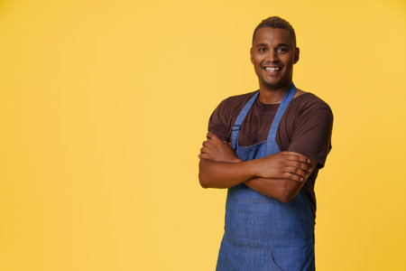 Cheerful African American cook wearing blue apron smiles in front of yellow background with copy space, suggesting concept of professional cook or chef in culinary or kitchen-related profession. High quality photoの写真素材