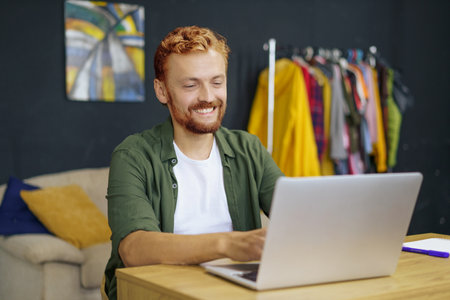 Caucasian man with red hair, sitting at desk in cozy home interior. He work on laptop, emphasizing concept of freelance work and ability to work from home. Man using his laptop to access his work remotely. High quality photoの写真素材