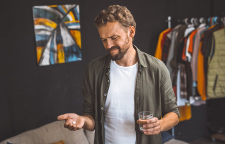 Man holding pill in one hand and glass of water in the other, isolated at home. Concepts of healthcare, wellness, and staying home for quarantine or isolation. The man is indoors and focused on taking his medication, while the glass of water provides a sense of refreshment and hydration. High quality photoの写真素材