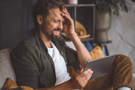 Man holding his head and covering his forehead with one hand while holding tablet pc in the other. He seems to be in discomfort and may be experiencing headache or other kind of pain. High quality photoの写真素材