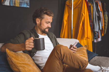 Man taking break at home, enjoying cup of coffee while focused on phone. Concept of spending time at home is emphasized, highlighting the importance of relaxation and leisure in our daily routines. High quality photoの写真素材