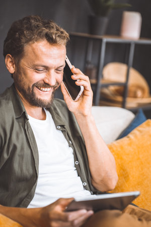 Happy man enjoying phone call while holding smartphone in one hand and tablet PC in other. He seat comfortably home, taking advantage of modern technology and convenience of being able to multitask. High quality photoの写真素材