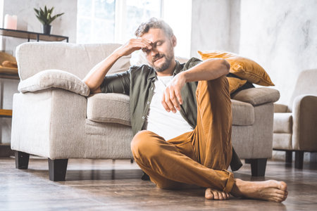 Middle-aged man resting on floor next to sofa home, moment of contemplation or crisis. Difficulties and challenges faced by men in society as they age, with focus on theme of middle age and its associated struggles. High quality photoの写真素材