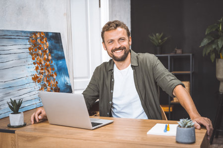Concept of remote work, freelance, and productivity in digital age. Cheerful and productive man, works from home, He smiling, focusing on his work, surrounded by digital technology and equipment. . High quality photoの写真素材