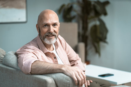 Happy mid-aged man with beard captured sitting on sofa in comfort of home. His infectious smile radiates warmth and joy, reflecting a sense of contentment. High quality photoの写真素材