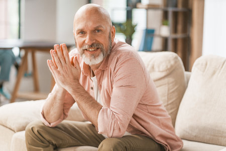 Smiling, happy senior man sitting on a sofa in the comfort of his home. With his hands held together, he radiates a sense of contentment and relaxation. . High quality photoの写真素材