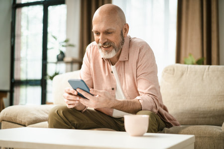 Happy mid aged man text message on phone while sit on sofa in home interior. High quality photoの写真素材