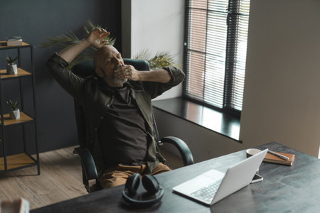 Tired senior man with silver beard yawns while working remotely from comfort of home. Realities of fatigue and exhaustion that can accompany remote work. . High quality photoの写真素材