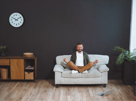 Young, handsome man in relaxed pose on sofa, practicing lotus asana with closed eyes and serene smile on face. He surrounded by tools of online education, with notebook displaying online lessons nearby on the floor. High quality photoの写真素材