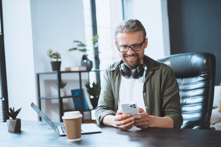 European man is taking break in office and using phone to send text message, illustrating concept of easy communication. Casual and relaxed work environment. High quality photoの写真素材
