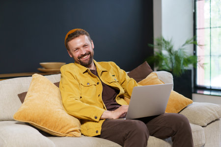 Happy and productive work-from-home scenario. Young man is seen sitting comfortably on sofa, smiling contentedly as he works on his laptop. High quality photoの写真素材