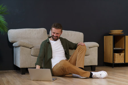 Man sitting on floor near sofa in house and working on laptop. He is freelancer or remote worker, using technology and internet to do job. High quality photoの写真素材