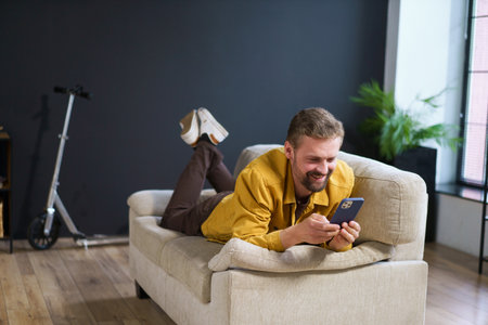 Smiling man enjoying time at home, sitting on sofa with phone. He appears handsome and content engages in texting or messaging on mobile device. High quality photoの写真素材