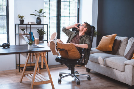 Businessman taking break in office and having conversation on phone while adopting relaxed posture. This image represents the idea of communication and relaxation in a professional setting. High quality photoの写真素材