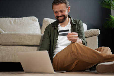 Happy, smiling customer sitting on floor next to sofa while shopping online using credit card for online auction. Concept of online payment systems, e-commerce, and online shopping in modern and technological setting. High quality photoの写真素材