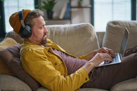 Young man in casual clothing sitting on sofa home, playing online game on laptop. He is fully concentrated on game, wearing headphones and using the keyboard to control action on screen. High quality photoの写真素材