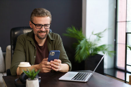 Positive freelancer who working from comfort of home. Smiling man seen texting message on phone while taking well-deserved break from work, resting near notebook. . High quality photoの写真素材