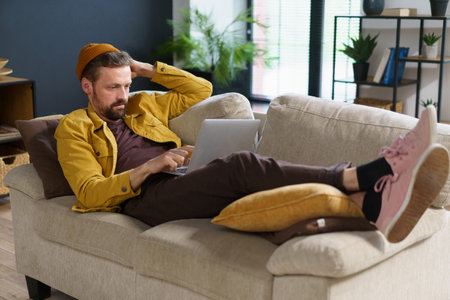 Wise programmer deep in thought, contemplating how to write code for program. Freelancer comfortably laying on sofa while working from home, embracing flexibility and benefits of remote work. . High quality photoの写真素材