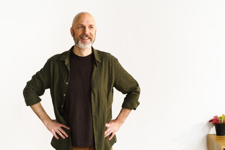 Smiling mid-aged bald man with silver beard standing confidently in front of white wall. His...
