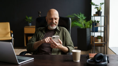 Bald and respectable man with gray beard. He captured in moment of joy, smiles while reading messages on phone. Computer, cup of coffee, and audio headphones are present on the table. High quality photoの写真素材