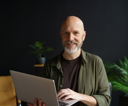 Happy senior man with warm smile, holding laptop in hands home. Mans cheerful and joyful expression, highlighting his contentment and happiness. . High quality photoの写真素材