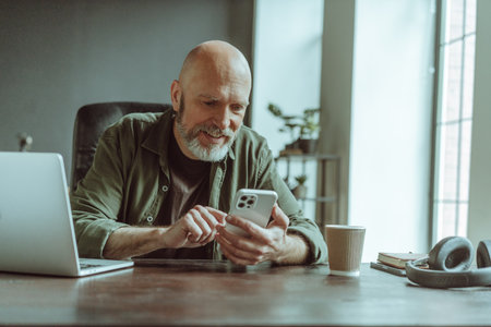 Mature old man comfortably seated home, actively engaged in texting message on phone. Desk adorned with laptop, headphones, and cup of coffee, indicating setup conducive to work and relaxation. . High quality photoの写真素材