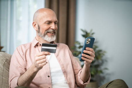 Senior man holds credit card in one hand and mobile phone in other. Convenience and accessibility of digital transactions for elderly, emphasizing their ability to adapt to modern technology. . High quality photoの写真素材