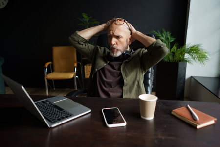 Man sitting in chair in loft office with unpleasant surprise. His face shows mixture of shock and disbelief, and he screaming with hands raised in air. Desk holding a laptop and other office supplies. High quality photoの写真素材