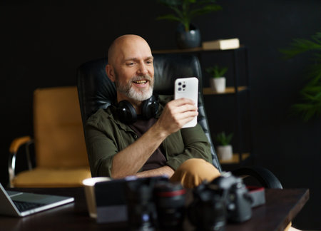 Mid aged man with bald head and silver beard engrossed in reading news on mobile phone. With laptop placed on desk in background, image captures essence of connected and digitally-oriented lifestyle. . High quality photoの写真素材
