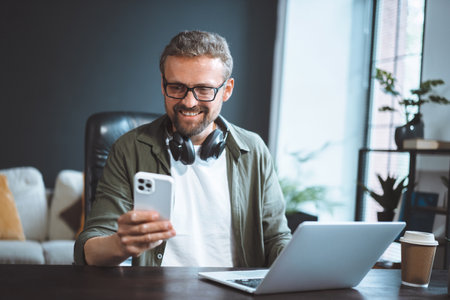 Happy and smiling man is sitting in a loft office, dressed in casual wear, working on his laptop while texting a message on his phone. High quality photoの写真素材