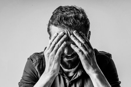 Black and white photo of a man looking extremely sad and depressed. He sitting on white background and has face covered with both hands, showing the despair and hopelessness he feels. High quality photoの写真素材