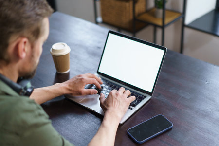 man working on laptop in loft office. man seated at desk with laptop, mobile phone, and coffee paper cup placed on it. typical workspace setup, giving viewer sense of modern work environment. High quality photoの写真素材
