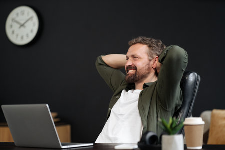 Happy and smiling man taking break and relaxing in office chair. He working on laptop, while enjoying cup of coffee. Modern and productive workplace, with a focus on technology and communication. High quality photoの写真素材