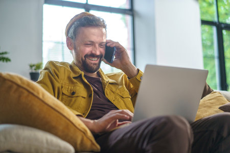 Smiling man having conversation on phone while working on laptop on sofa in office. He dressed in casual wear and most likely programmer or IT professional. High quality photoの写真素材