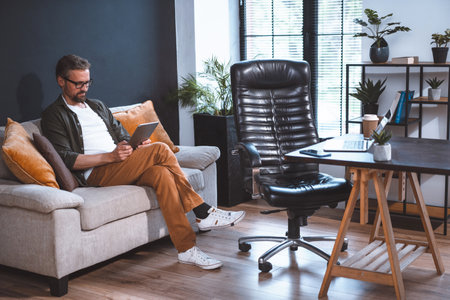 Freelancer sitting on sofa, looking relaxed as he uses tablet PC. Laptop on desk and empty chair can be seen nearby in room. Concept of relaxation after work, and ease and comfort of working from home. High quality photoの写真素材