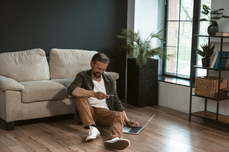 Mid-aged man is holding credit card and using laptop to make online payment. He entering payment information on website, which can not be seen on computer screen. High quality photoの写真素材