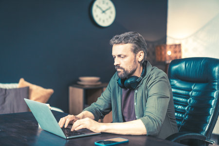 Man sitting at desk home, typing on laptop and chatting online through messaging app. He dressed in casual clothing and surrounded by technology, with wireless mobile phone router visible in the background. This image is perfect for representing remote work, telecommuting, or the modern workplace. High quality photoの写真素材