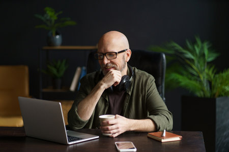 Smiling senior man at home, engrossed in reading news on laptop while holding cup of drink. Enjoying moment of relaxation and staying informed in comfort of his own space. High quality photoの写真素材