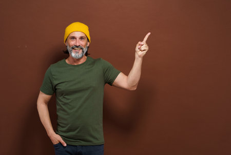 Smiling European man in a yellow hat points his hand to the side with copy space on a brown background. Product placing, advertising concept. High quality photoの写真素材