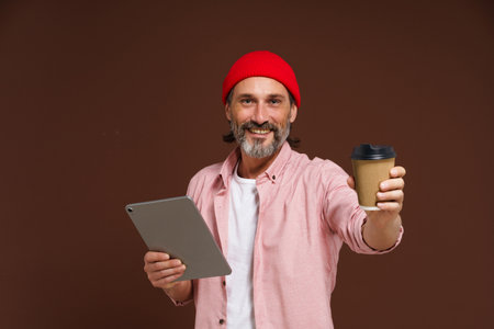 Work and coffee concept. Smiling mature man in casual clothing holds tablet in hand while offering craft cup with coffee in other hand. Happy mature freelancer caucasian man in red hat isolated on brown background. High quality photoの写真素材