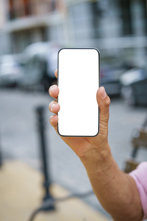 Close-up of mans hand holding mobile phone with white screen display, offering ample copy space. Background features urban cityscape, providing modern and vibrant backdrop. . High quality photoの写真素材