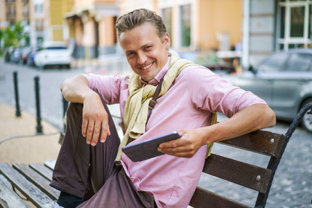 Smiling man sitting on bench in city, and using tablet PC. With gadget in hand, he effortlessly stays connected to digital world, browsing internet, engaging social media, or accessing various digital content. . High quality photoの写真素材
