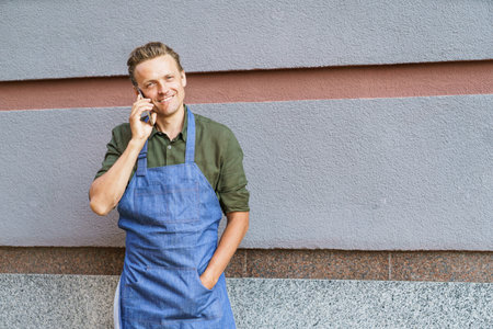 Concept of service readiness and the willingness to assist customers. Worker in blue apron, holding mobile phone, and ready to take order from client against wall background. High quality photoの写真素材