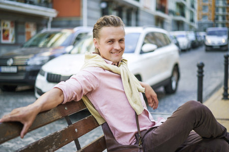 Happy and handsome individual with lazy and carefree expression, smiling while sitting on bench in urban city. Persons relaxed demeanor reflects their contentment and enjoyment in moment. . High quality photoの写真素材