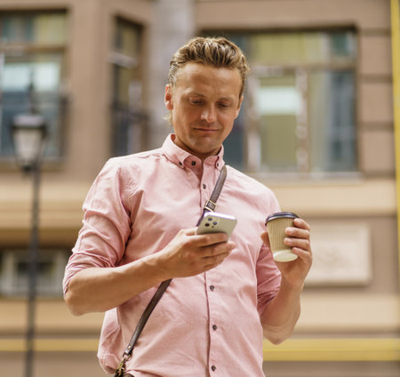 Mid-aged man texting message on phone. He enjoy cup of coffee from paper cup on summer city street. Intersection of technology, as man engages in mobile communication while enjoying coffee break. . High quality photoの写真素材