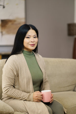 Cute Asian MILF sitting on couch with cup of coffee home. The image captures moment of relaxation and leisure as woman enjoys her coffee break in comfort of her own home. . High quality photoの写真素材